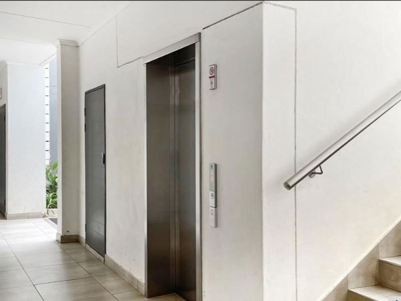 A clean, well-maintained apartment building hallway featuring a stainless steel elevator, staircase with a metal handrail, and neutral tiled flooring.