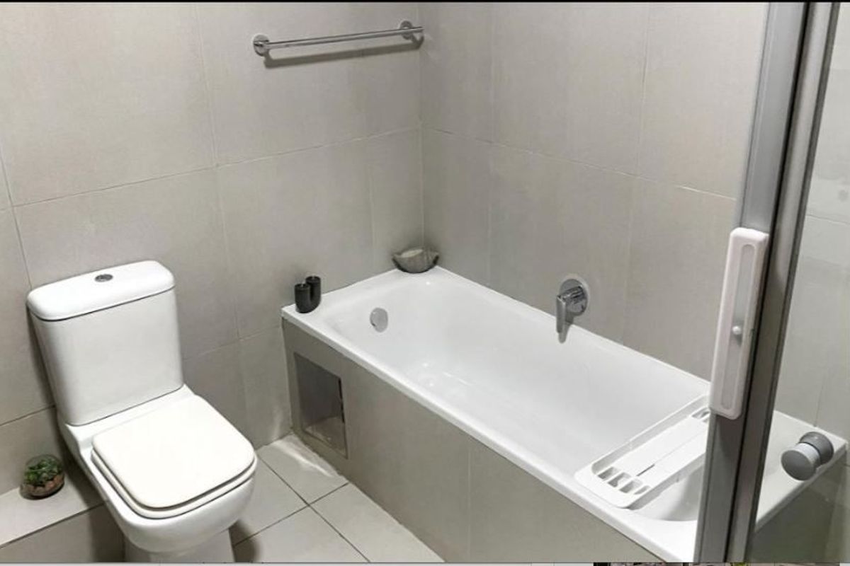 A clean, neutral-toned bathroom featuring a built-in bathtub, modern toilet, and full-height wall tiling with a glass shower screen.