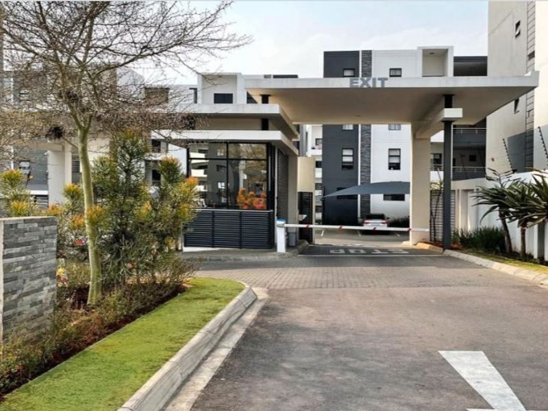 Modern apartment complex entrance featuring a secure gatehouse, boom gate access control, paved driveway, and well-maintained landscaped surroundings.