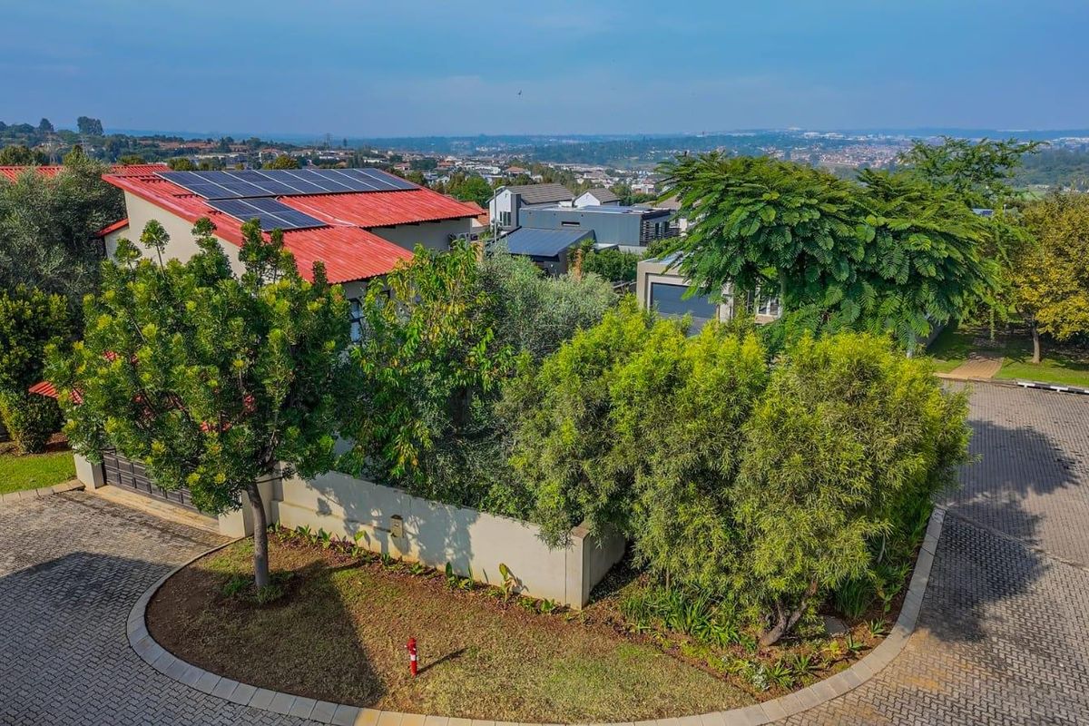 Aerial view of a residential property featuring a metal roof with integrated solar panels, surrounded by established landscaping and paved driveway access.