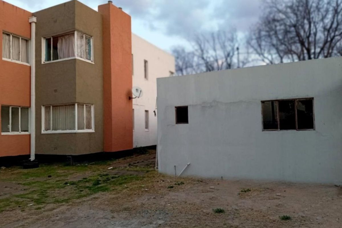 Rear view of a multi-story residential building and a detached single-story outbuilding with visible exterior wear and unlandscaped ground area.
