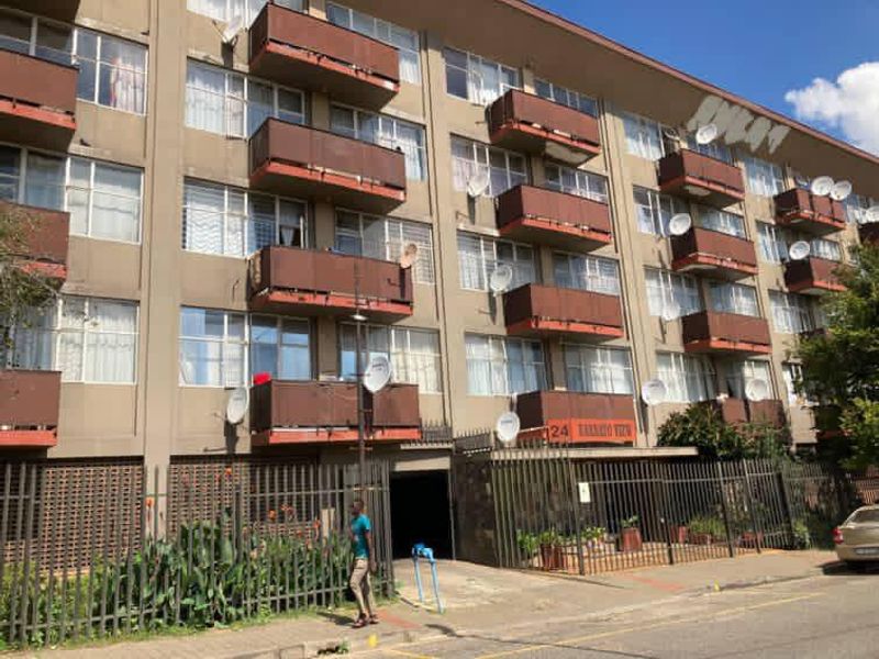 Street-level view of a multi-story apartment building featuring multiple balconies, security fencing, and exterior facade wear.