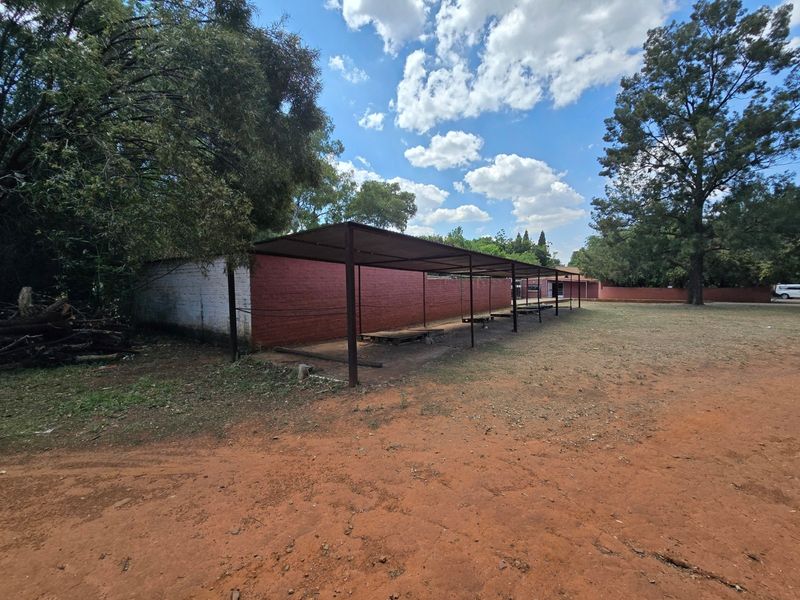An outdoor area featuring a long metal carport structure and a painted brick boundary wall set within a large, unpaved garden space.