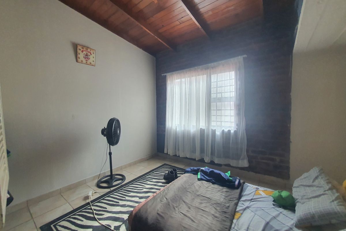 Bedroom featuring an exposed timber beam ceiling, a rustic exposed brick feature wall, tiled flooring, and a window fitted with security bars.