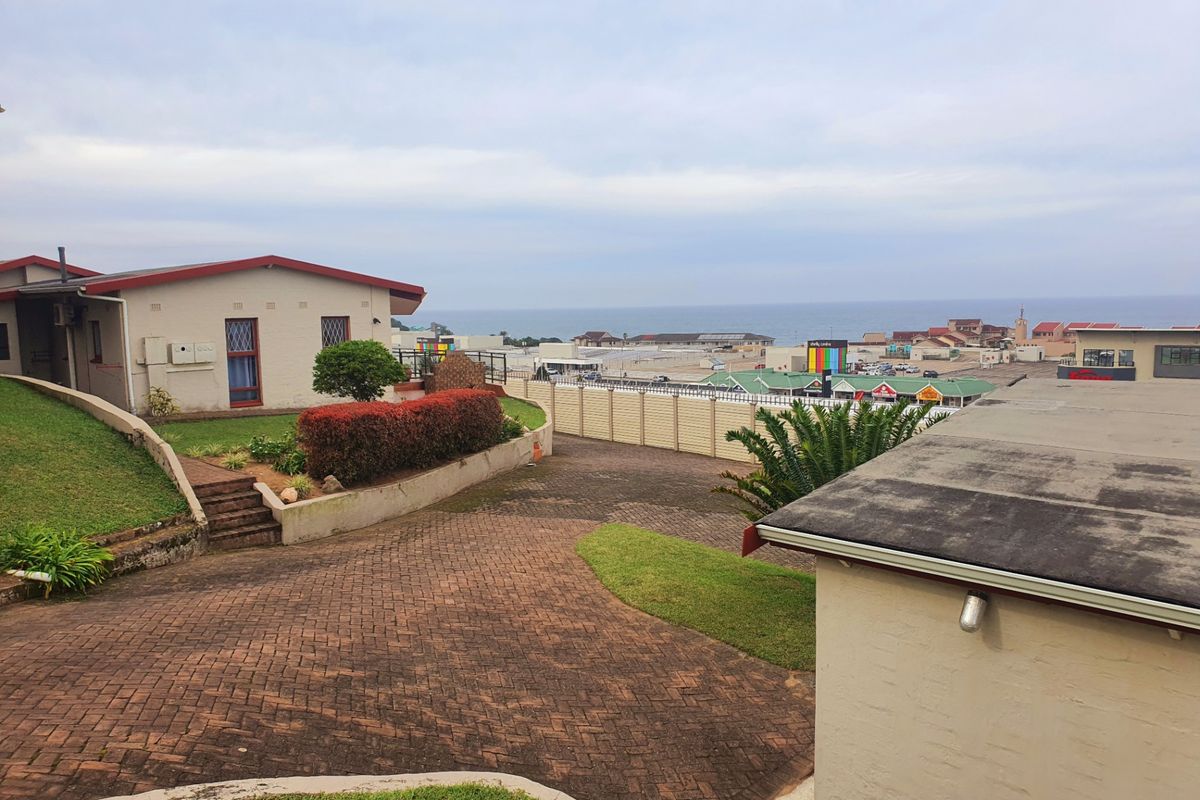 An elevated view of the property exterior featuring a paved driveway, retaining walls, and a boundary fence with a distant coastal outlook.