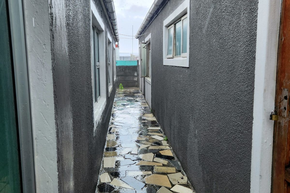 A narrow side passage between two exterior walls featuring paved flooring and guttering systems.