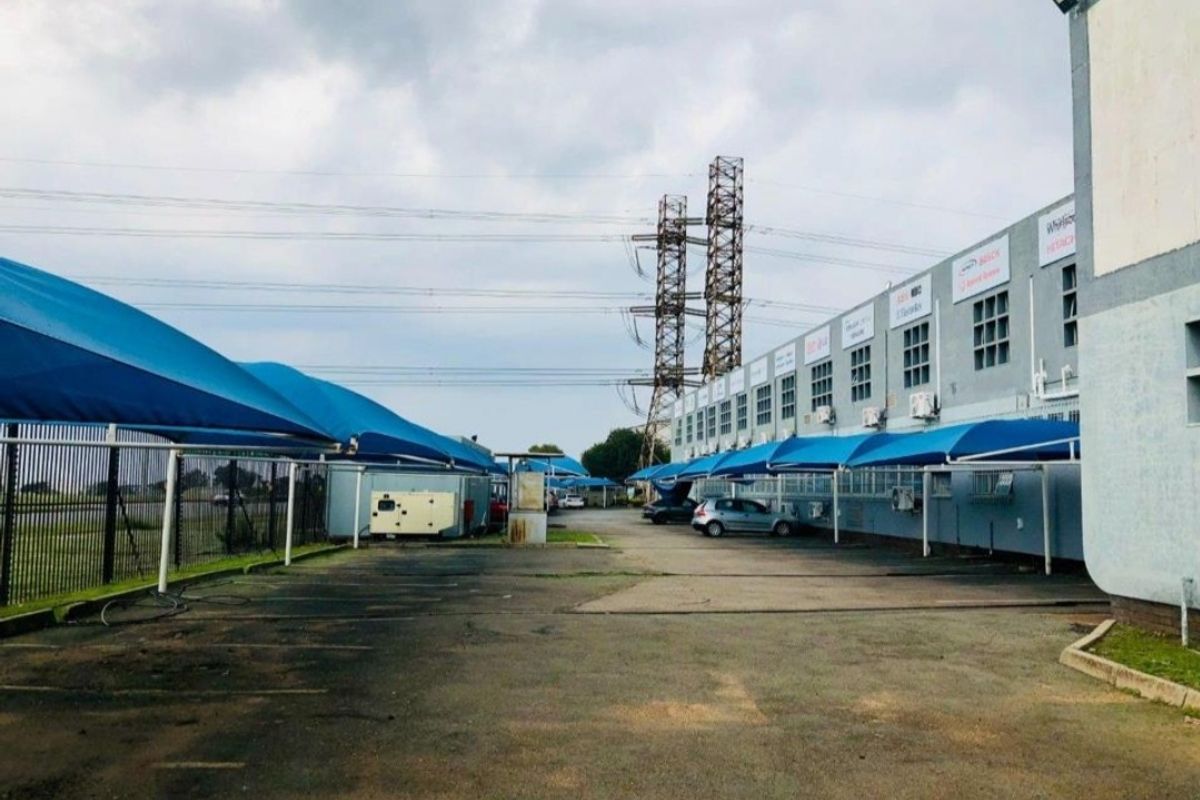 Industrial property exterior featuring an asphalt parking area with shade cloth carports and wall-mounted air conditioning units along the building facade.