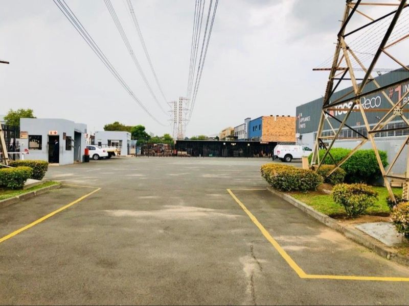 Industrial property entrance featuring a paved driveway, security guard house, and perimeter fencing with high voltage power lines overhead.