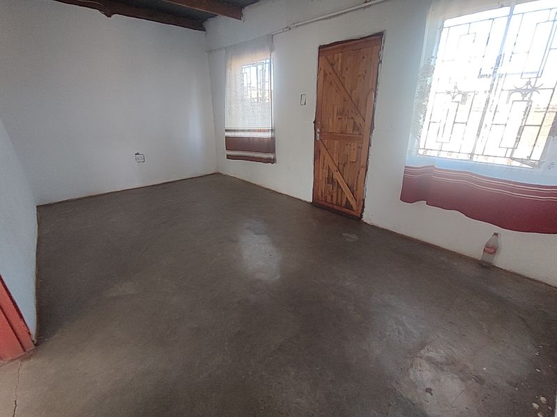 An empty bedroom featuring exposed timber ceiling beams, security bars on windows, and a raw cement floor finish.