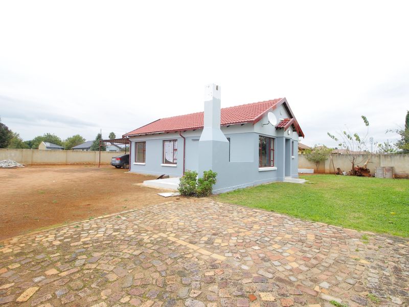A detached single-story house featuring a tiled roof, external chimney, and a cobblestone driveway, set on a spacious plot with a perimeter wall.