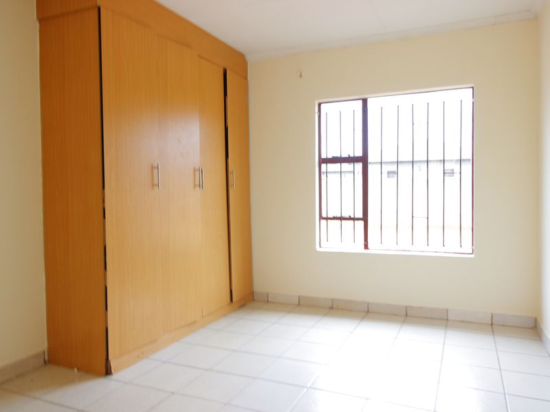 A vacant bedroom featuring tiled flooring, built-in wooden cupboards, and a window fitted with security bars.