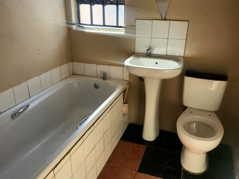 A functional bathroom featuring a bathtub, pedestal basin, and toilet, with tiled finishes and security bars on the window.