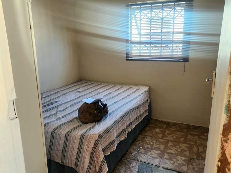 A compact bedroom featuring a window with security bars and blinds, requiring significant flooring repairs and cosmetic updates to improve the overall condition.