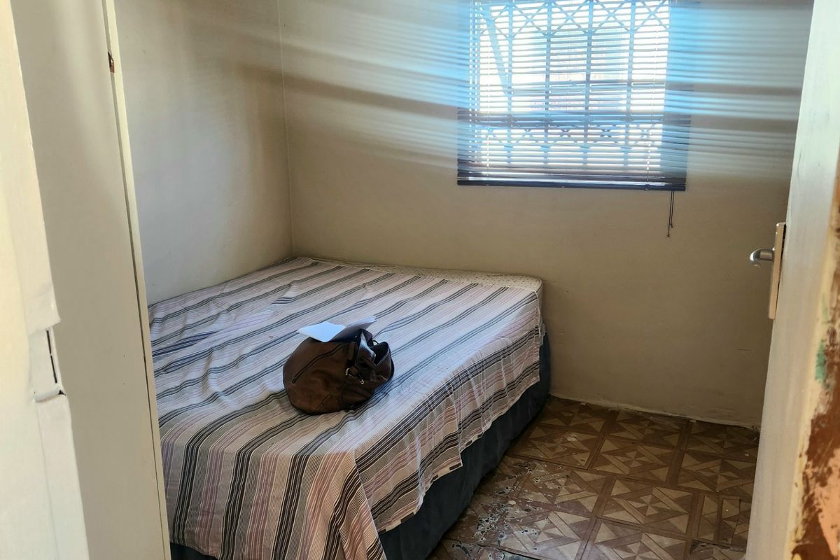 A compact bedroom featuring a window with security bars and blinds, requiring significant flooring repairs and cosmetic updates to improve the overall condition.