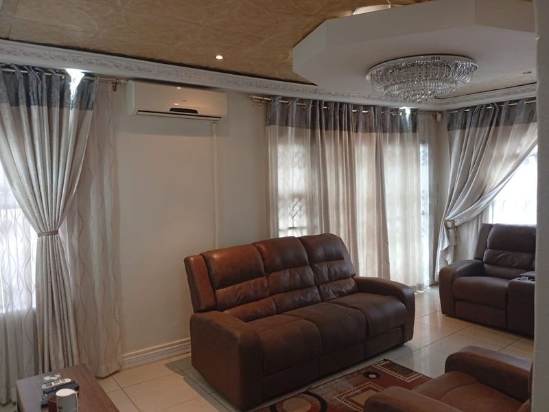 A well-maintained living room featuring tiled flooring, decorative ceiling molding, a central crystal chandelier, and a wall-mounted split-system air conditioner.