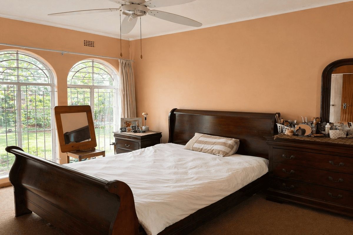 A spacious bedroom featuring arched windows with security bars, a ceiling fan, and neutral carpet flooring.