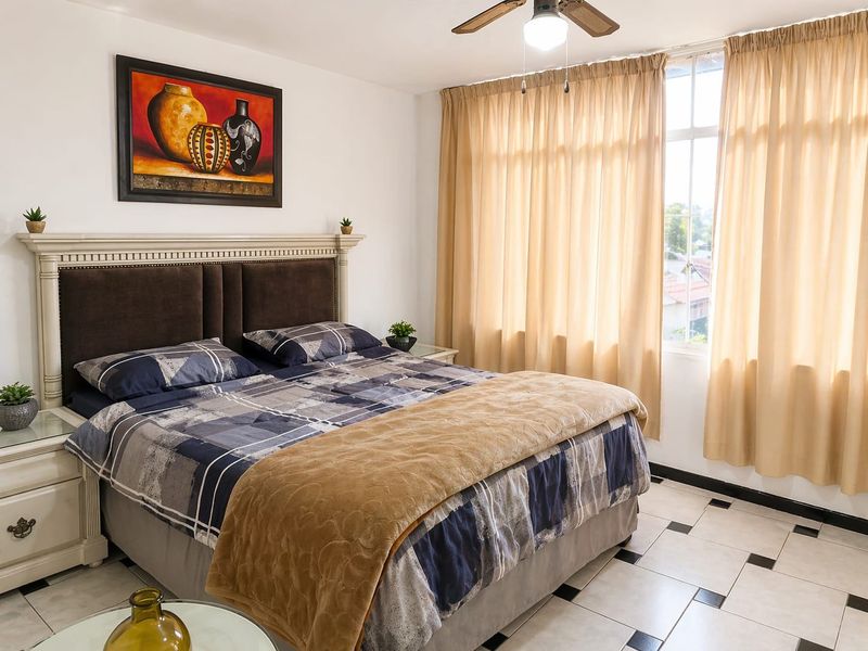 A well-lit bedroom featuring tiled flooring, a ceiling fan, and large windows with curtains, providing a comfortable and functional living space.