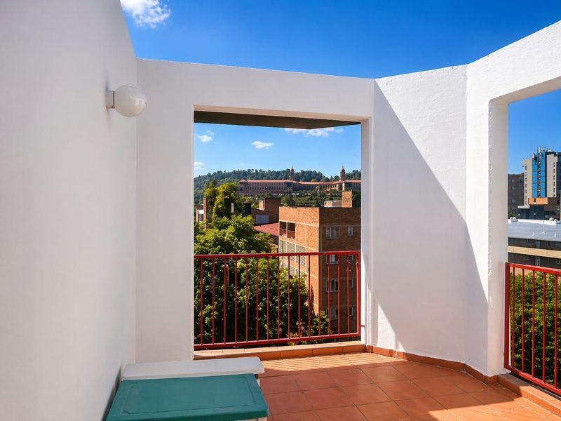 A bright, open-air balcony featuring terracotta floor tiles, white rendered walls, and red metal railings, offering an elevated urban view.