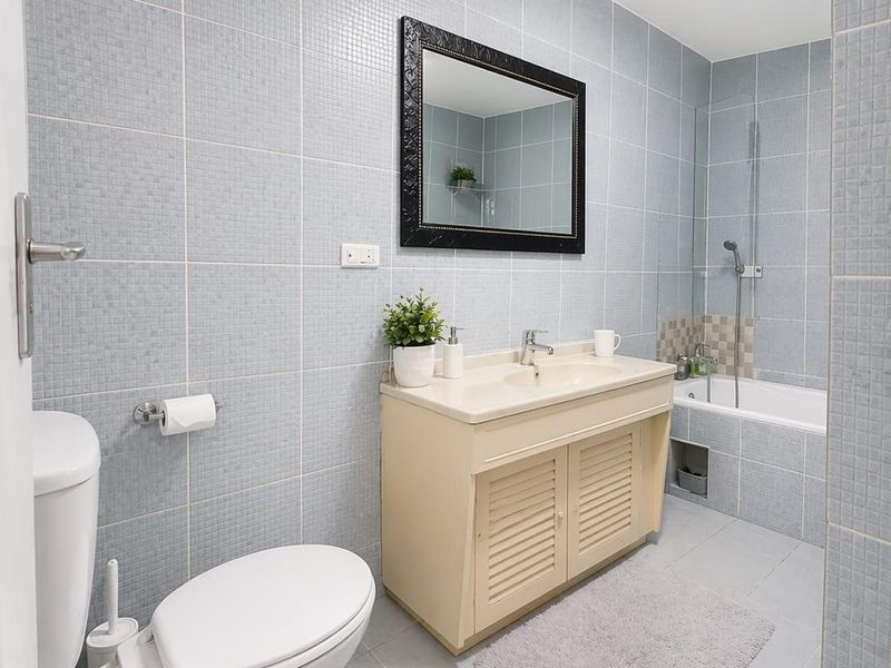 A functional bathroom featuring full-height blue mosaic wall tiling, a built-in bathtub with shower mixer, and a vanity cabinet with integrated basin.