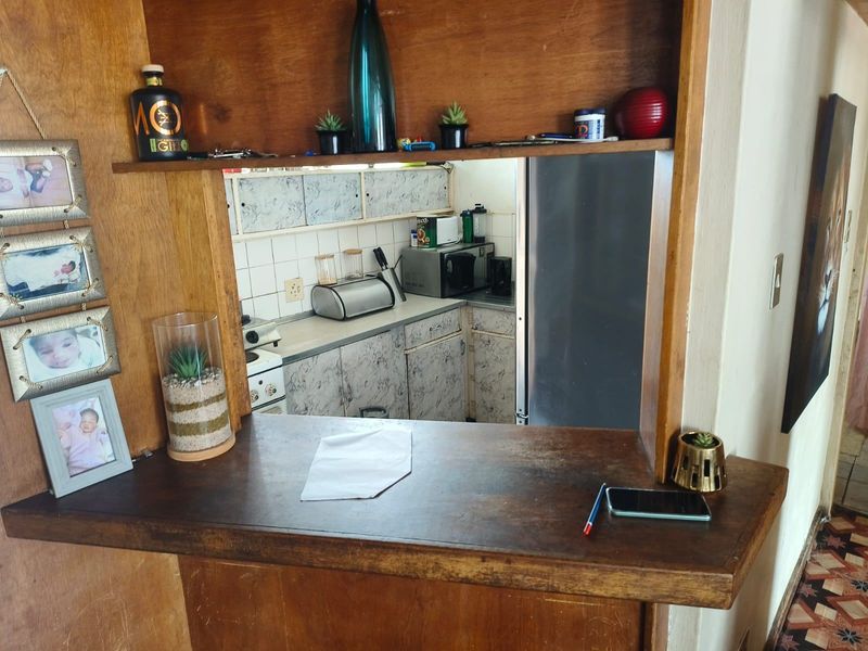 A view through a wooden breakfast bar into a dated kitchen featuring built-in cabinetry and a tiled backsplash.