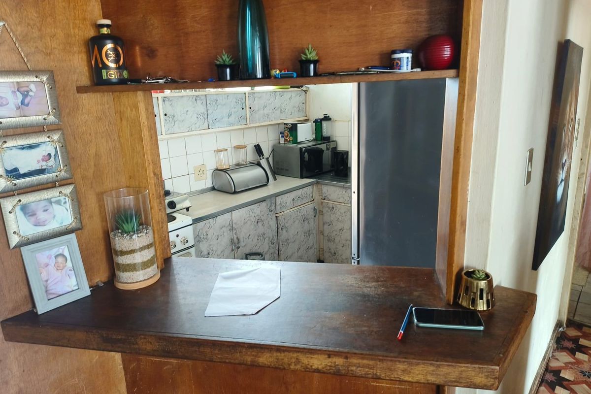 A view through a wooden breakfast bar into a dated kitchen featuring built-in cabinetry and a tiled backsplash.