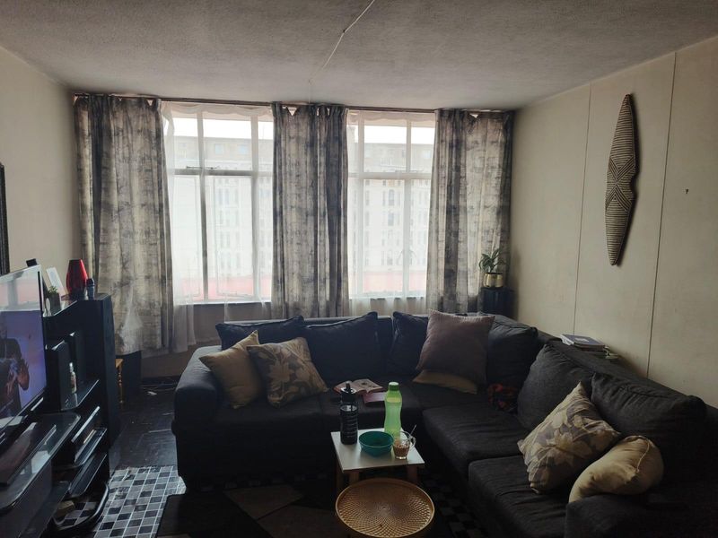 A living room featuring large windows that provide natural light, with tiled flooring and potential for cosmetic updates to the walls and ceiling.