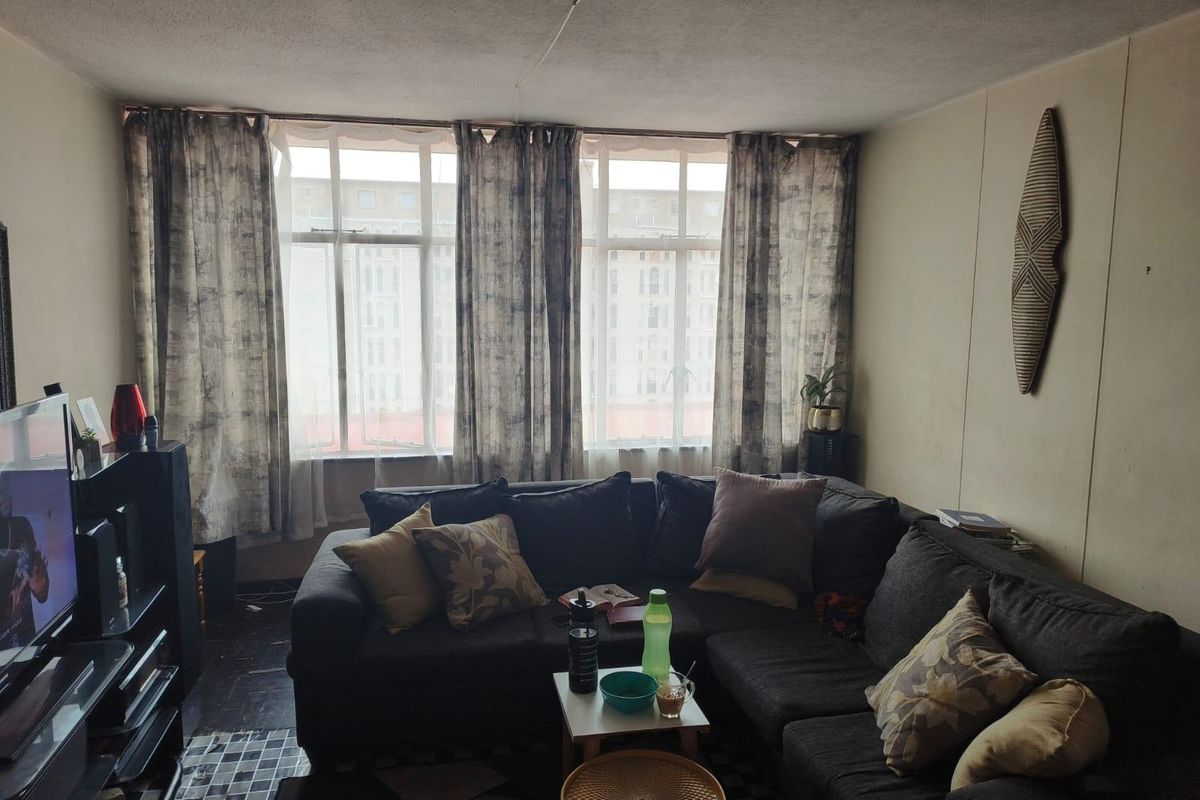 A living room featuring large windows that provide natural light, with tiled flooring and potential for cosmetic updates to the walls and ceiling.