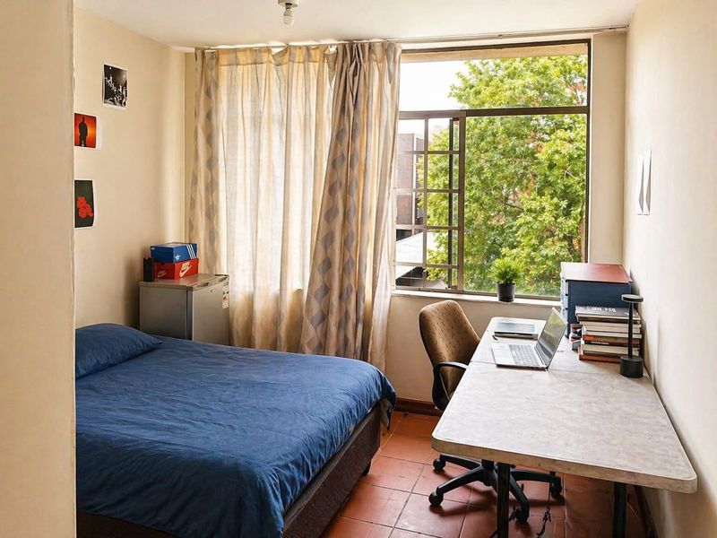 A functional bedroom featuring terracotta tile flooring and a large window that provides natural light and a pleasant leafy outlook.