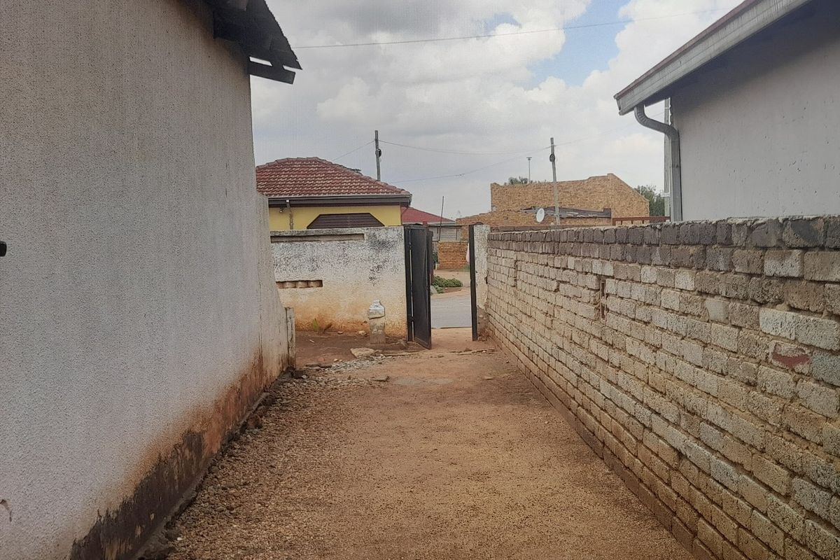 A narrow side passage between two structures featuring a brick boundary wall and an access gate, showing signs of wear and weathering.