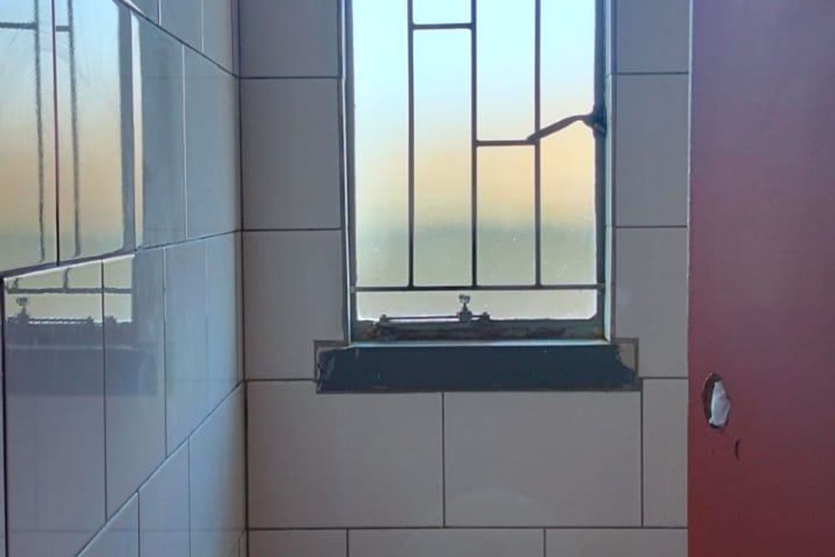 A compact bathroom featuring white wall tiles, a standard toilet, and a window with security bars.