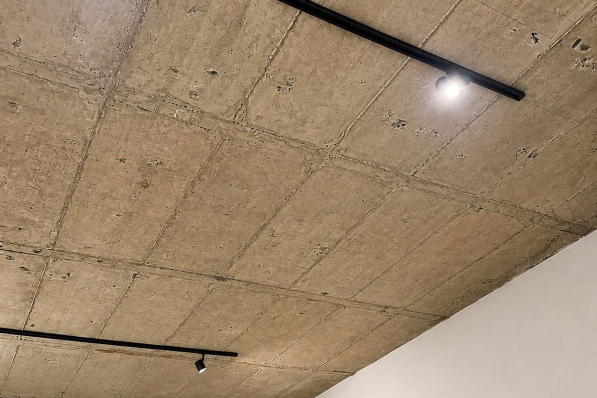 A modern bedroom interior featuring an exposed concrete ceiling with integrated track lighting and a wall-mounted split-system air conditioning unit.