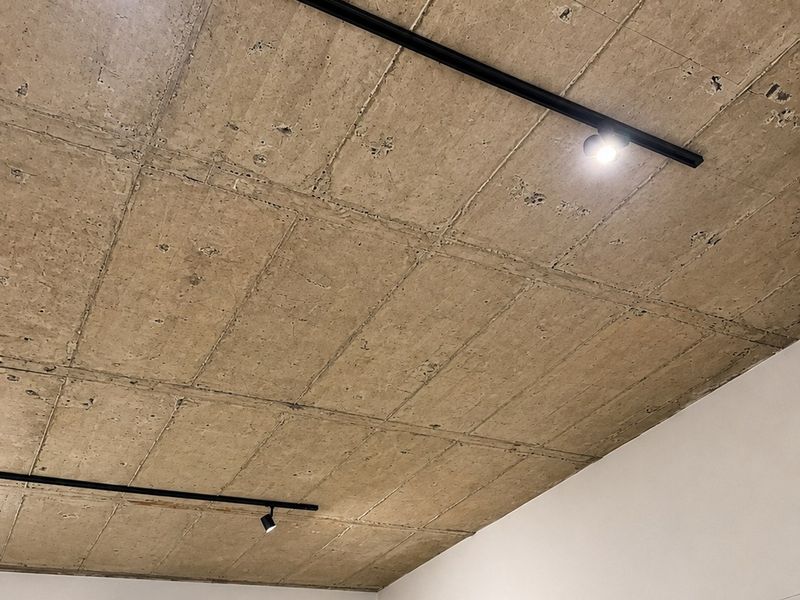 A modern bedroom interior featuring an exposed concrete ceiling with integrated track lighting and a wall-mounted split-system air conditioning unit.