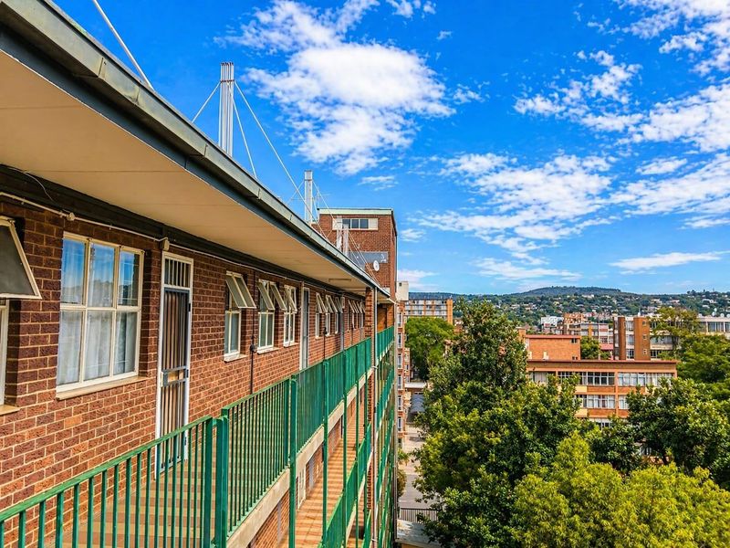 An exterior view of an apartment building balcony featuring brick masonry, green metal railings, and tiled flooring overlooking the surrounding neighborhood.