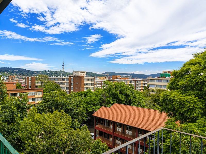 An elevated urban view overlooking surrounding apartment buildings, lush green trees, and the city skyline under a bright blue sky.