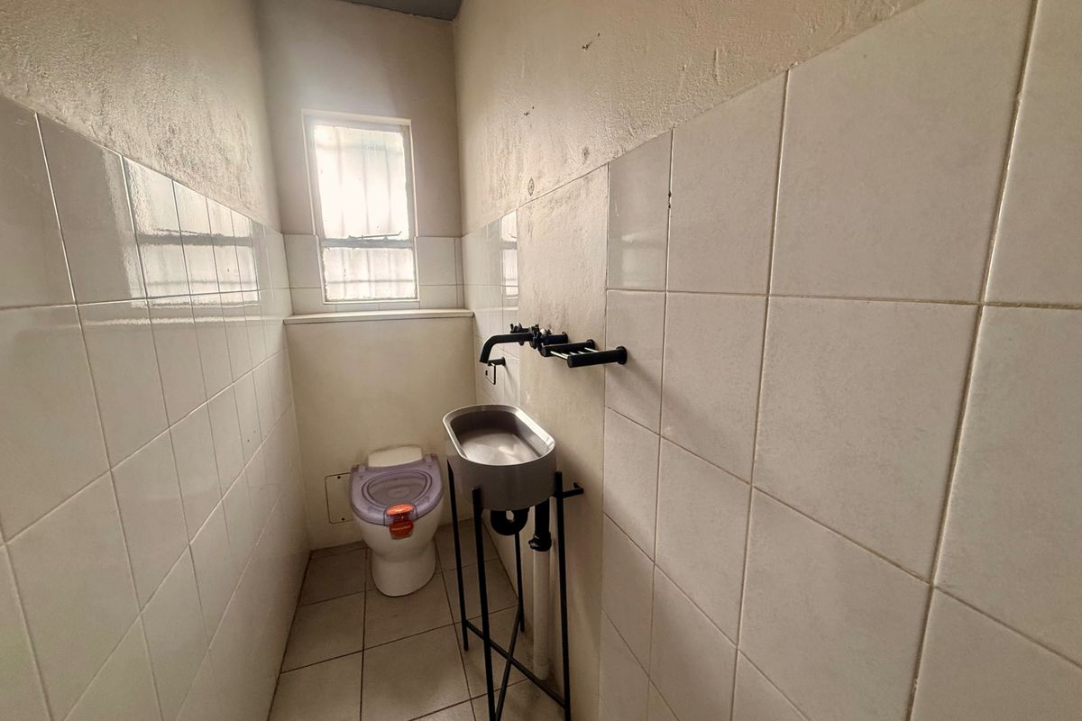 A compact commercial restroom featuring tiled walls, a stainless steel basin, and a window with security bars.