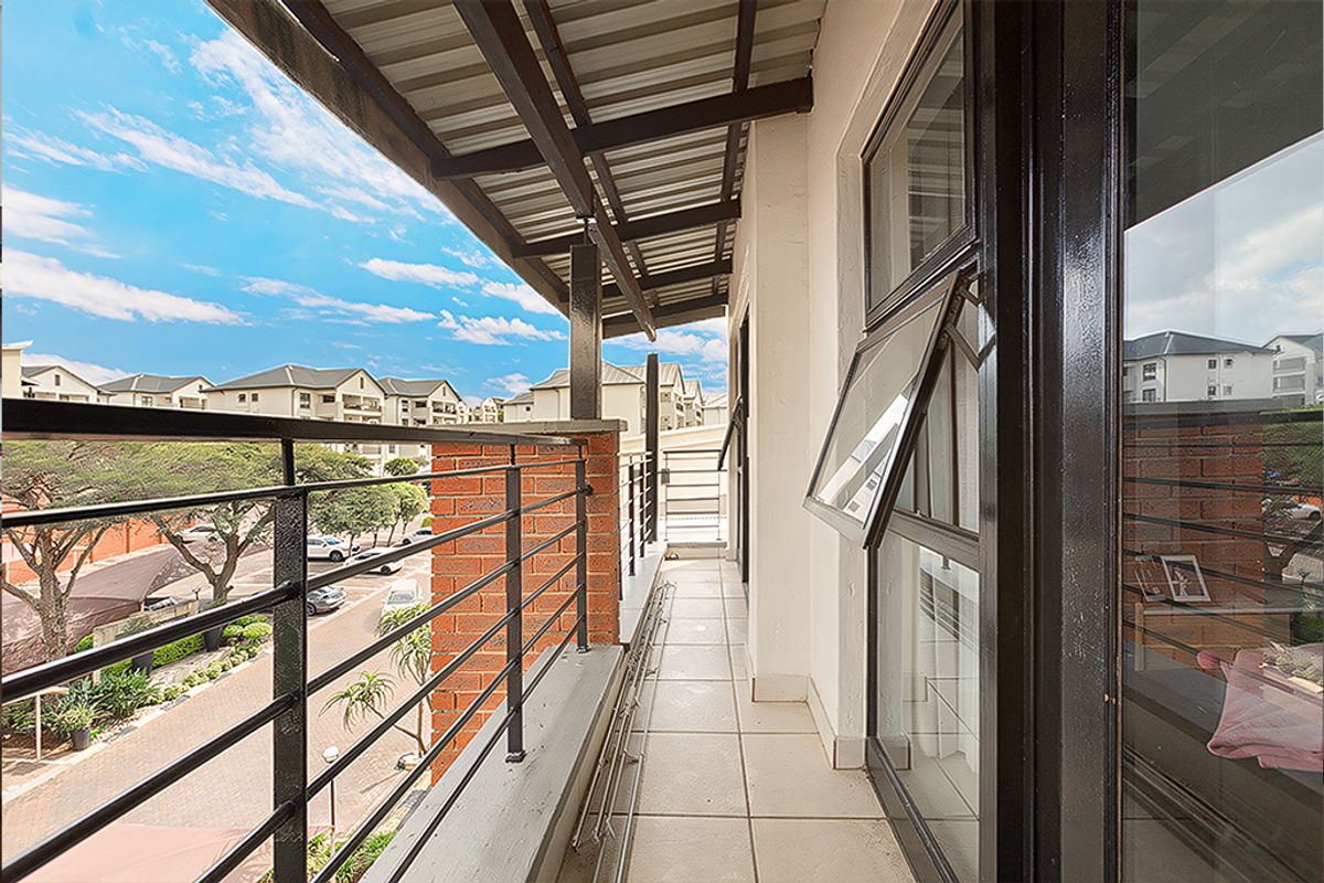 A well-maintained balcony featuring tiled flooring, a metal balustrade, and a covered roof, offering an elevated view of the surrounding apartment complex.