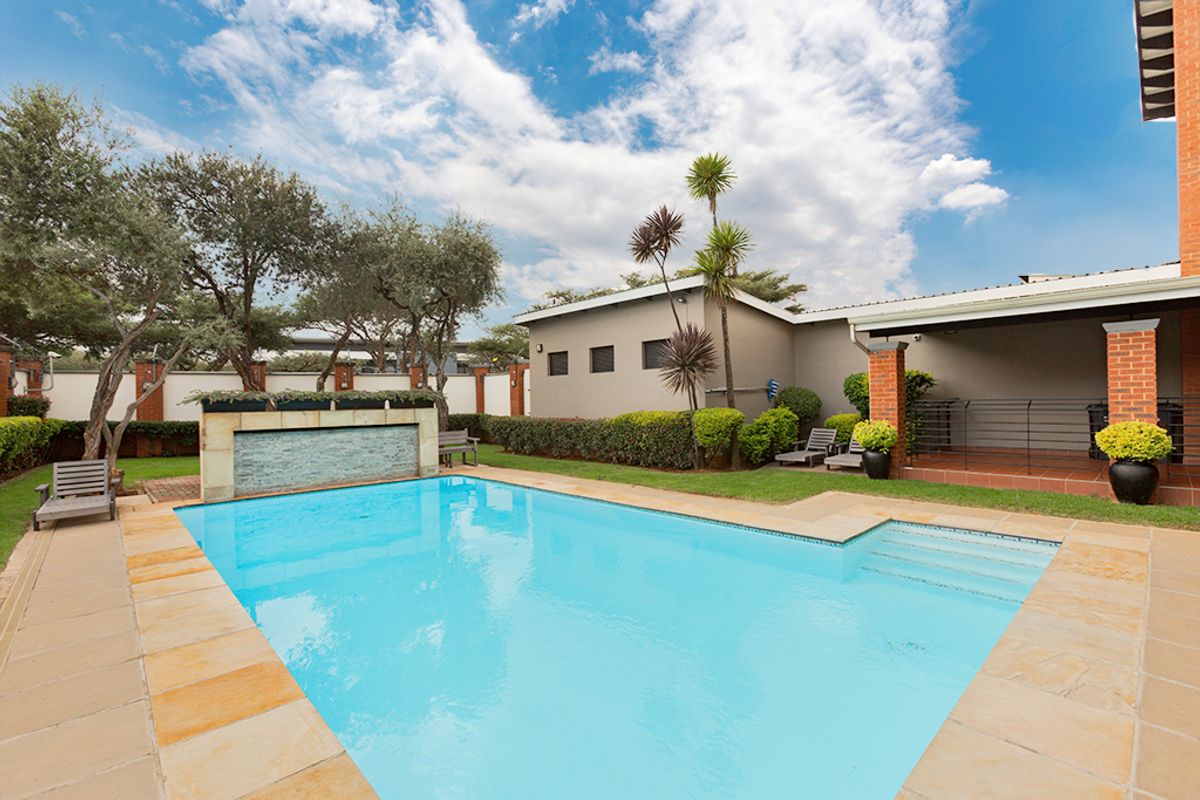 A well-maintained outdoor pool area featuring a stone-tiled pool deck, a decorative water feature, and an adjacent covered patio with brick structural elements.