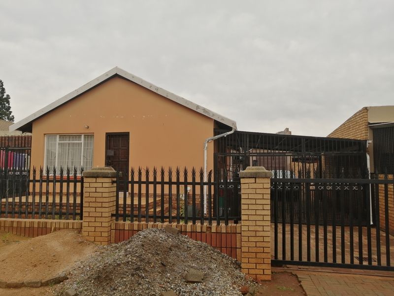 Front elevation of a single-story house featuring a brick perimeter wall, metal security fencing, and an attached carport.