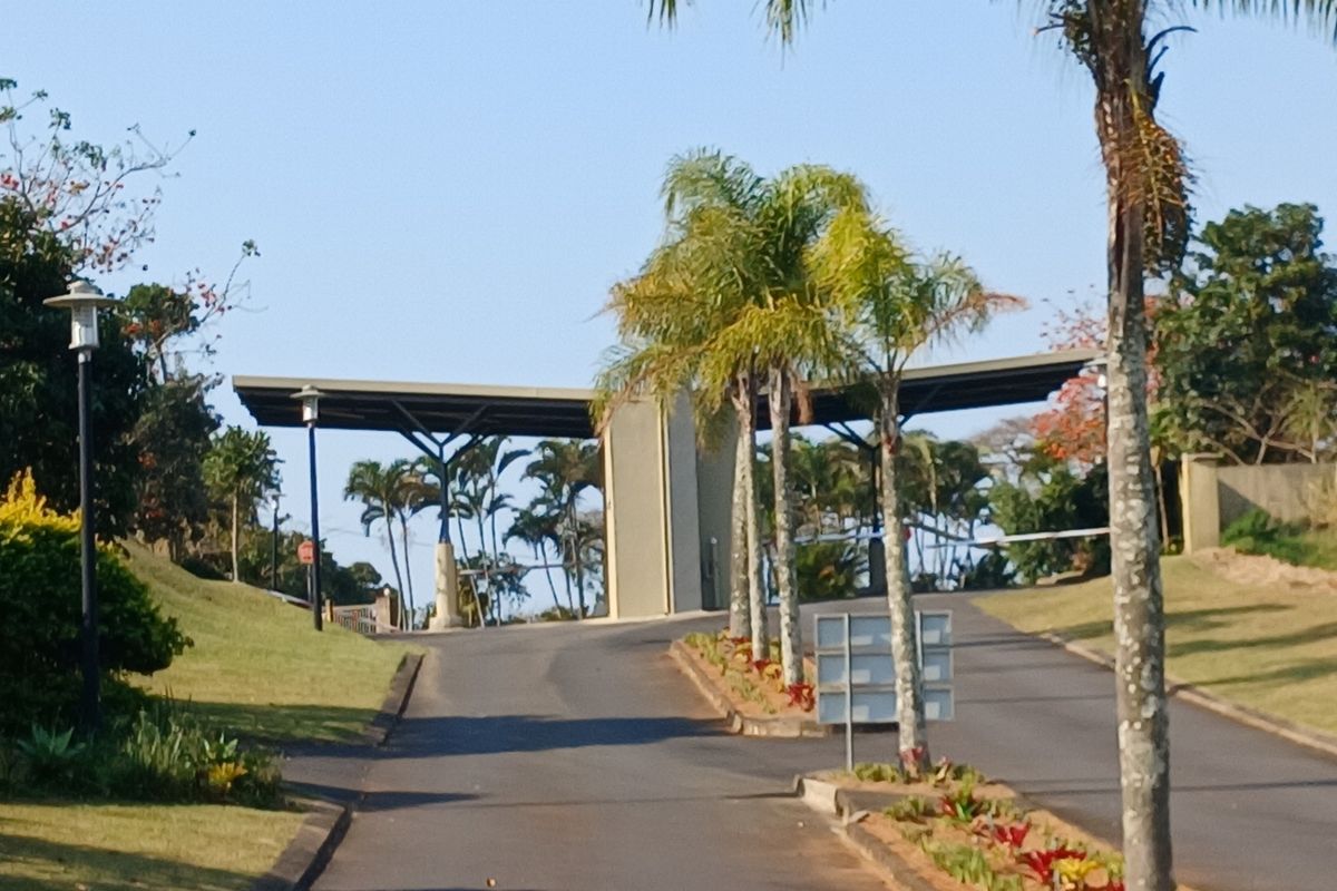 A view of the gated entrance to the residential estate, featuring a paved driveway, security boom gate structure, and landscaped palm trees.