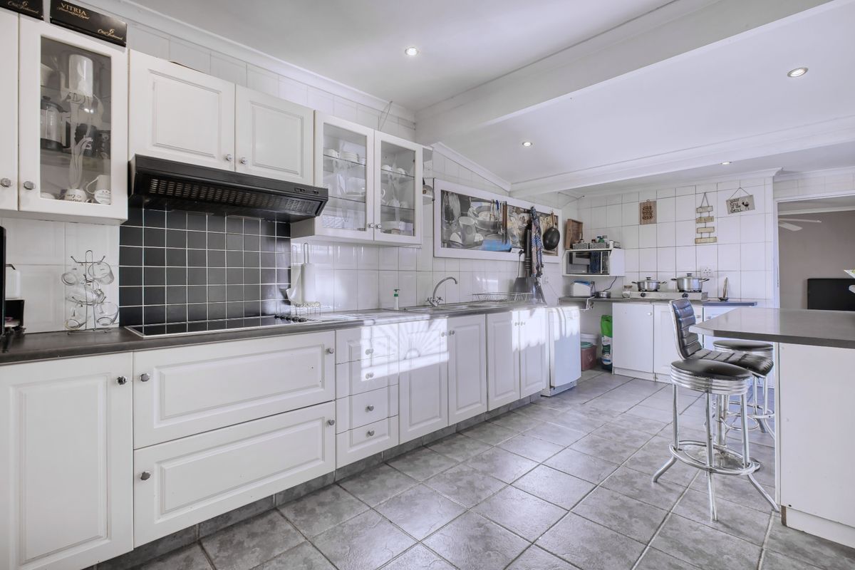 A functional kitchen featuring white cabinetry, black tiled splashback, integrated hob, extractor fan, and tiled flooring with a breakfast bar area.