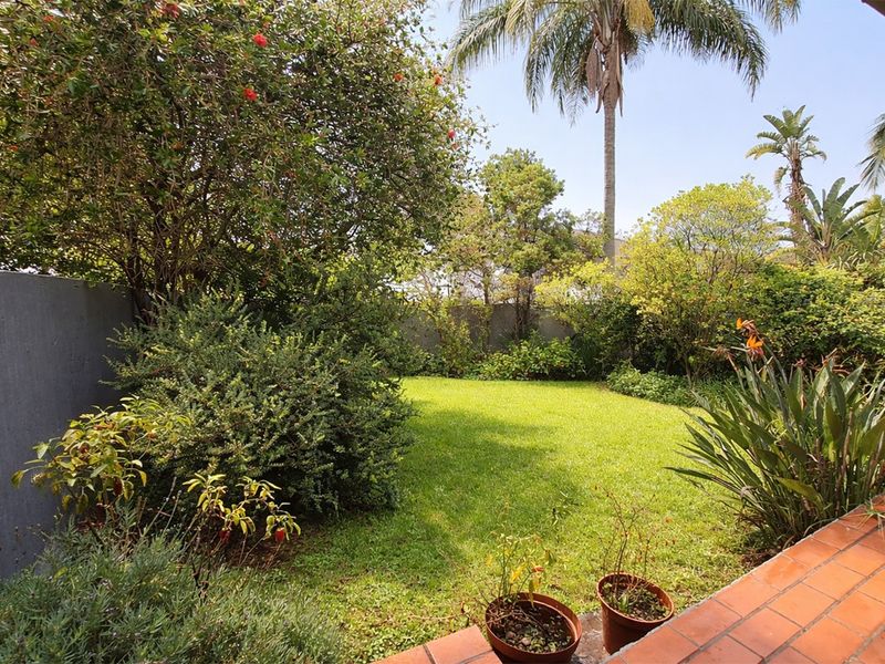 A private, well-maintained garden area featuring a lush lawn, mature trees, and a terracotta tiled patio border.