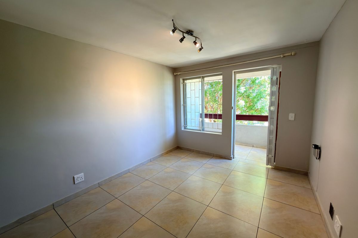 A bright, empty bedroom featuring tiled flooring, track lighting, and direct access to a private balcony through a glass door.