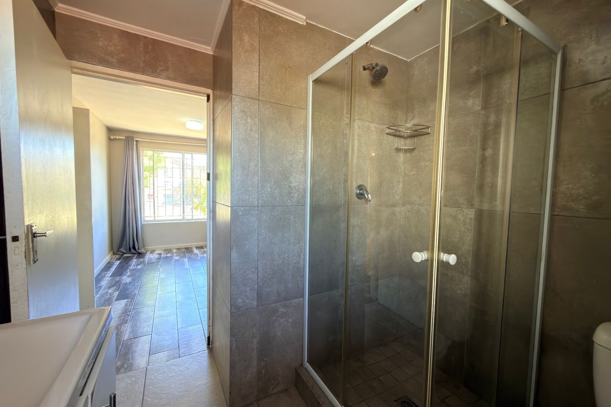 A clean bathroom featuring a glass-enclosed shower, neutral wall tiling, and a vanity basin, with an adjacent view into a bedroom area.