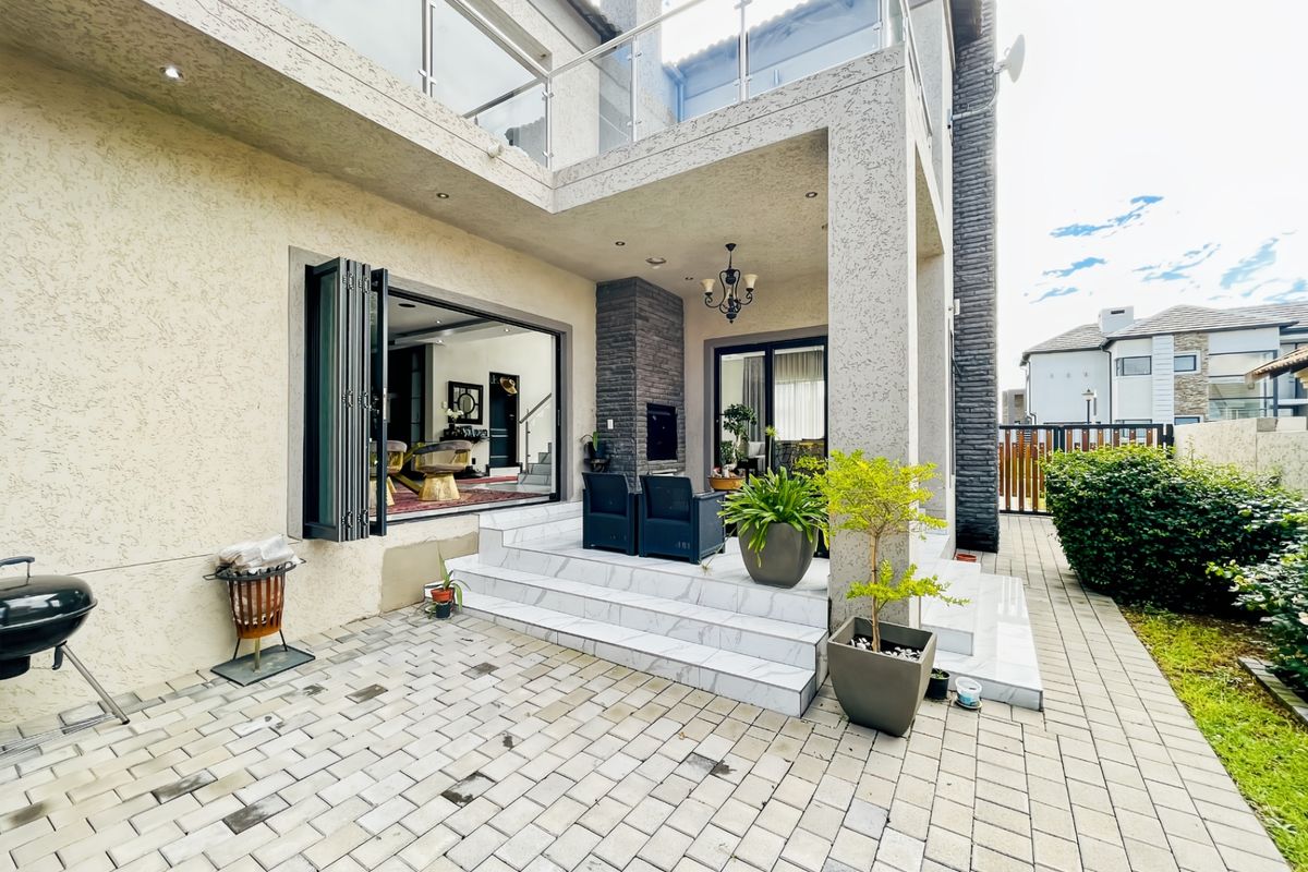A modern rear patio featuring stone cladding, a built-in fireplace, bi-fold doors leading to the interior, and a glass-balustraded balcony above.