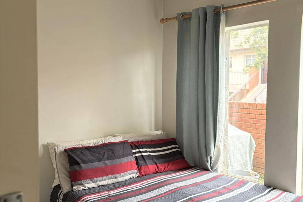 A compact bedroom featuring neutral wall tones and a window with a curtain rod, providing natural light and a view of the exterior.