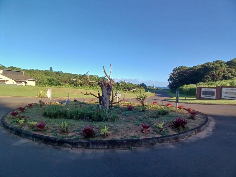 A view of a landscaped roundabout and entrance road within a residential estate, featuring established greenery and clear signage.