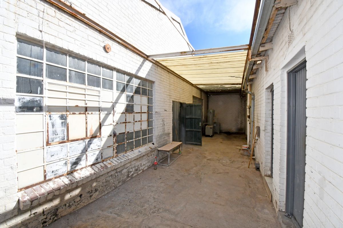 An industrial rear exterior area featuring weathered brick walls, steel-framed windows, and a covered walkway with corrugated roofing.