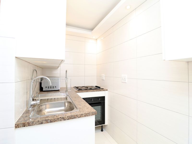 A compact, modern kitchen featuring white cabinetry, a stainless steel sink, built-in oven, electric hob, and clean tiled wall finishes.