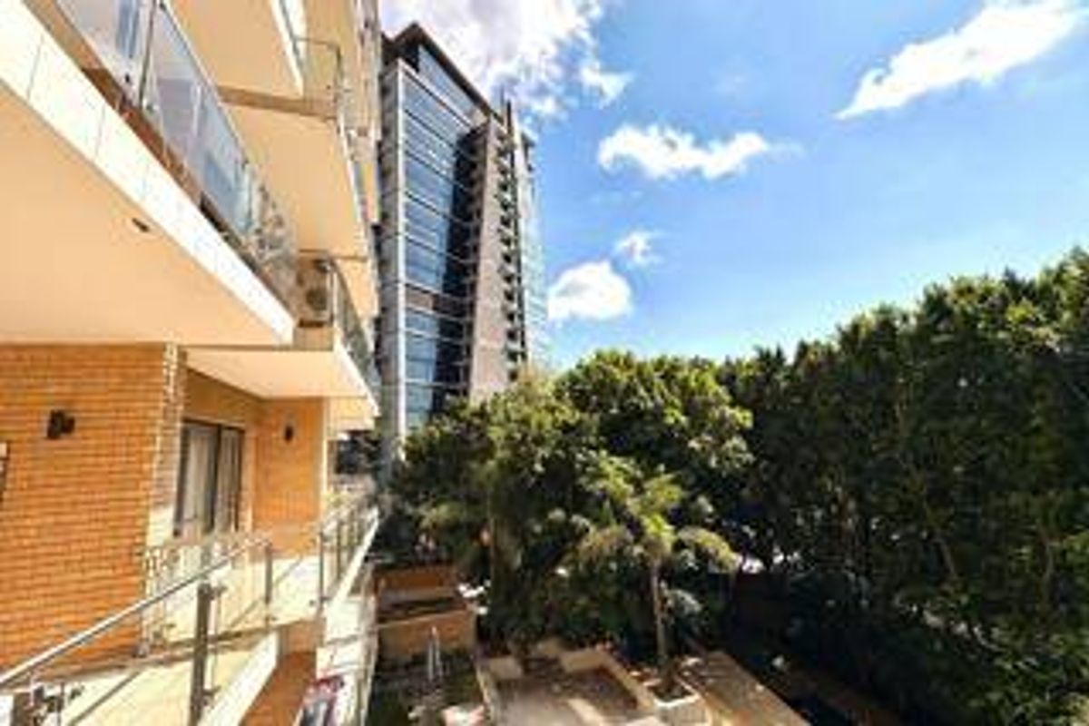 Exterior view of the apartment building featuring modern glass balustrades, brick cladding, and a view overlooking surrounding trees and neighboring high-rise architecture.