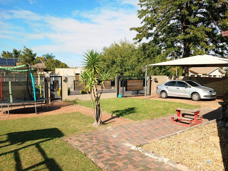 A well-maintained rear garden featuring a paved driveway, boundary wall with metal gates, and a shade sail structure over the parking area.